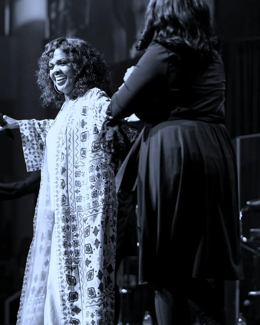 The artist laughing with a second vocalist on stage, microphone held out.