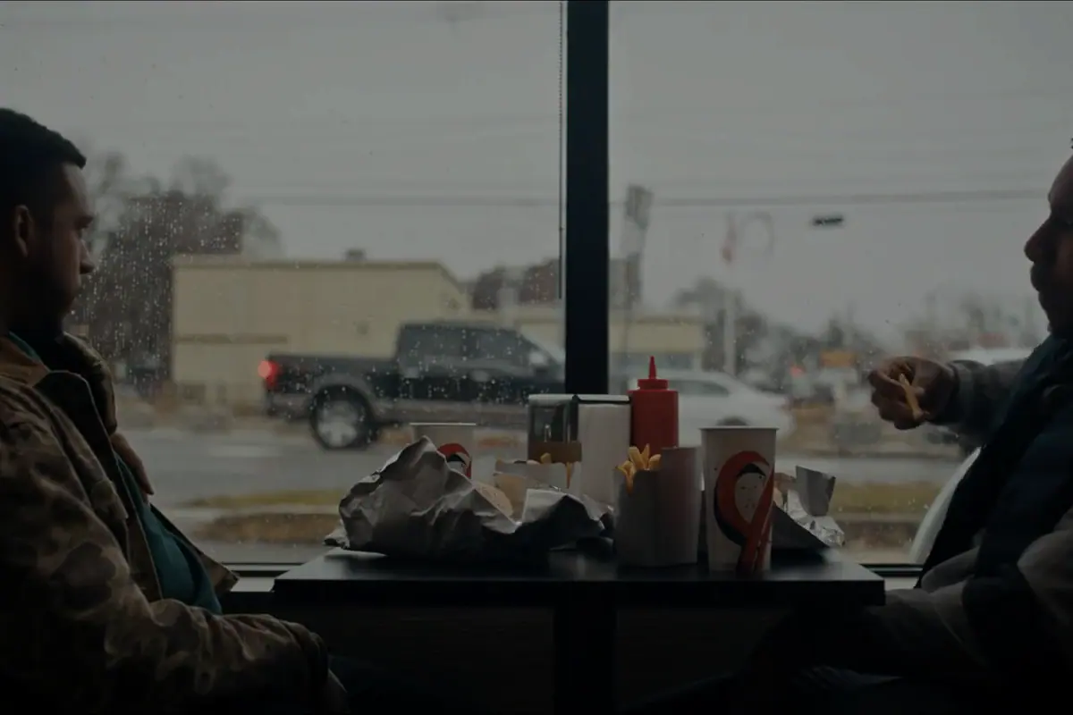 Two men talking across a diner counter, the parking lot through the window.